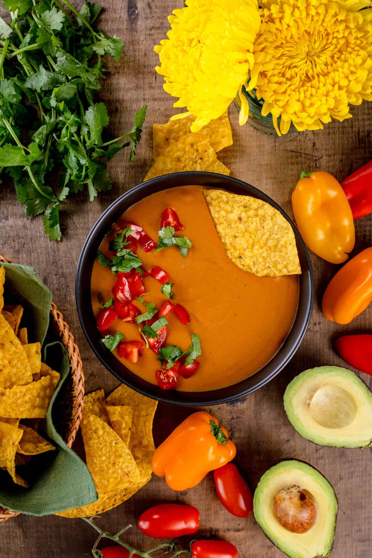 A big bowl filled with dairy free queso dip with fresh cilantro, tomatoes, and a chip on top. More chips, mini peppers, avocados, cilantro, and fresh flowers are next to the bowl on a wood tabletop.