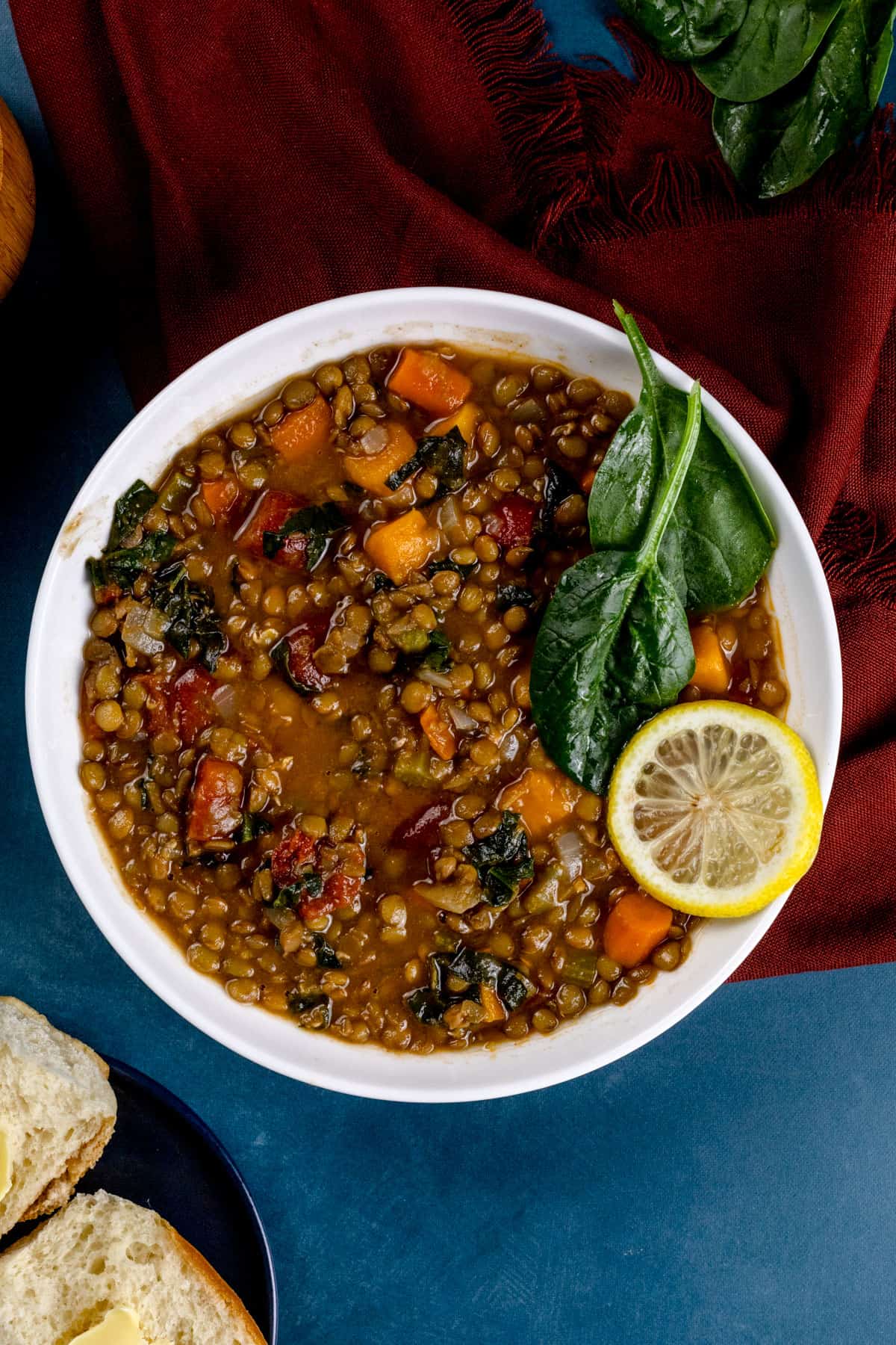 A big white bowl filled with lentil soup, more spinach, and a lemon slice. It rests on a red napkin. More fresh spinach and slices of bread are next to the bowl.