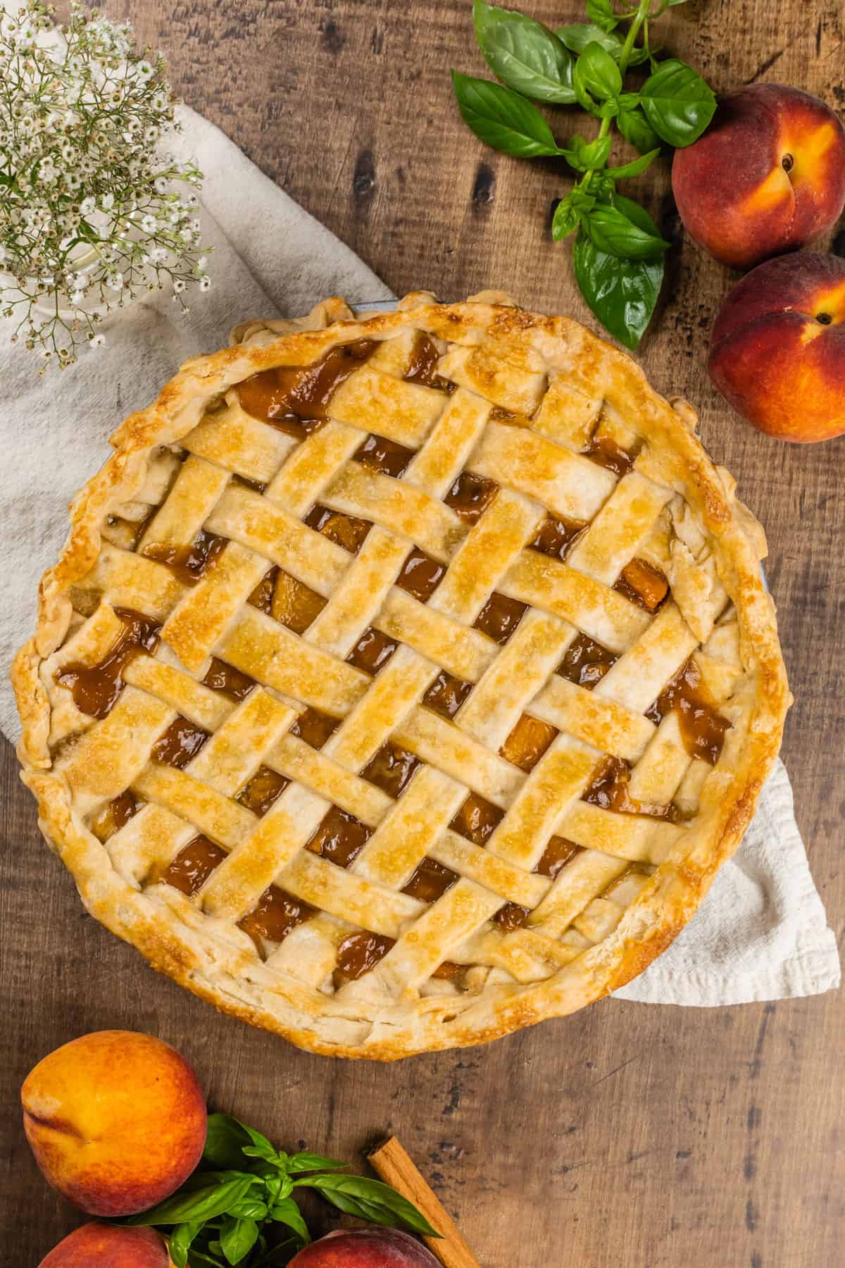 Finished peach pie with canned peaches. It has a lattice top crust and fresh peaches and herbs and flowers are besides the pie.