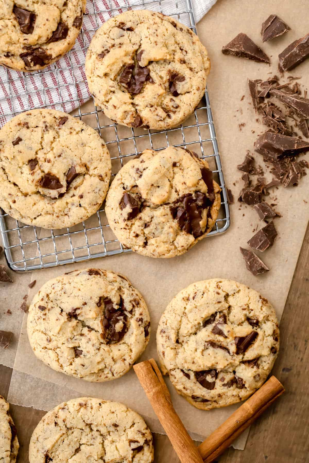 Many vegan chocolate chip cookies are on parchment paper and cooling racks with cinnamon sticks and chopped chocolate next to the cookies.