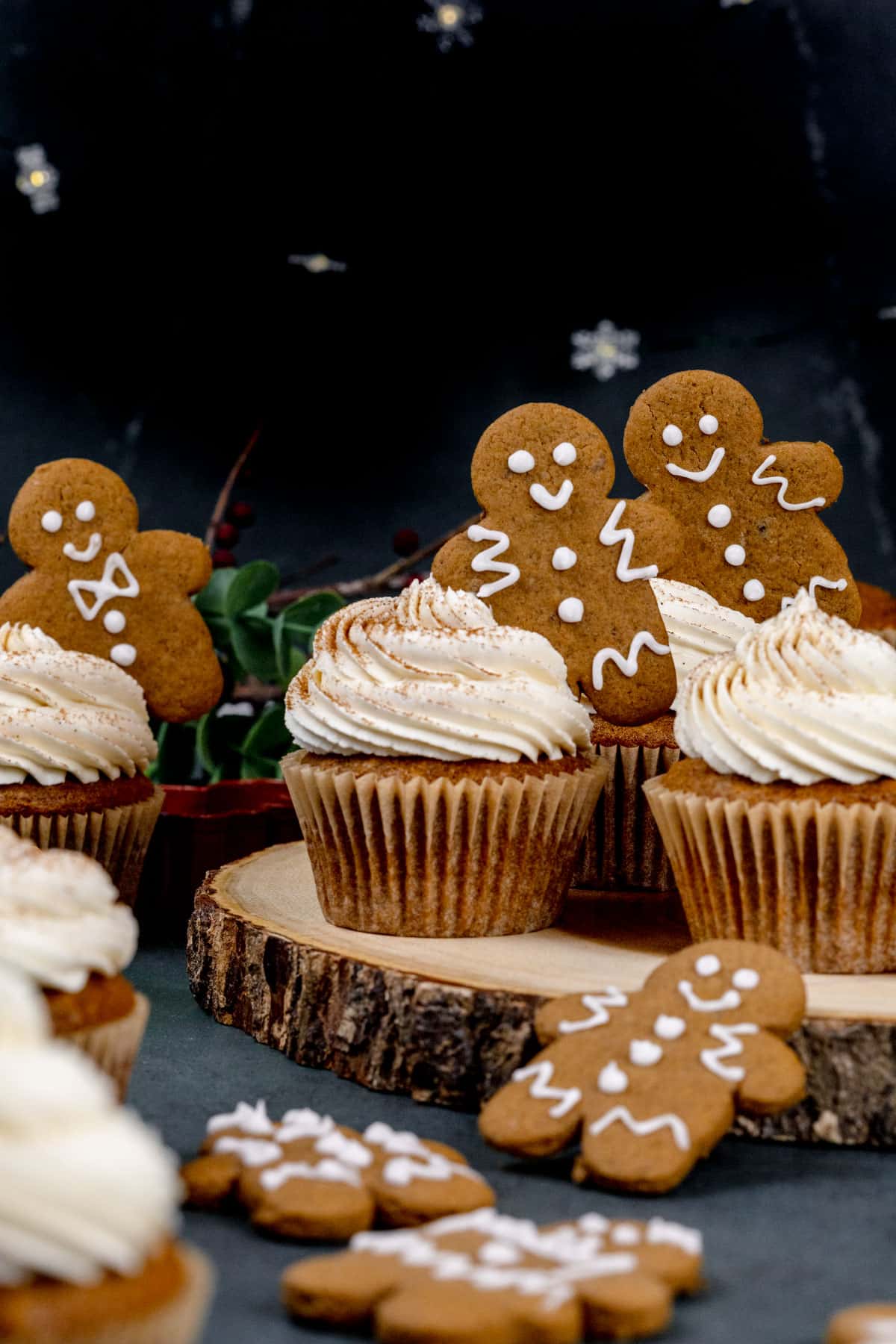 Multiple vegan gingerbread cupcakes on a table with swirls of frosting and tiny gingerbread men placed in each frosting swirl. More cookies are on the table surrounding the cupcakes.