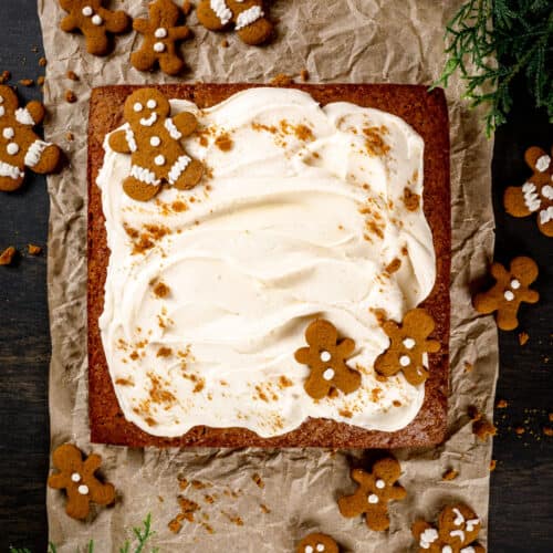A square vegan gingerbread snack cake on crinkled parchment paper on the table. Swirls of white frosting decorate the top of the cake and tiny gingerbread men cookies are on top of the cake and all around it.