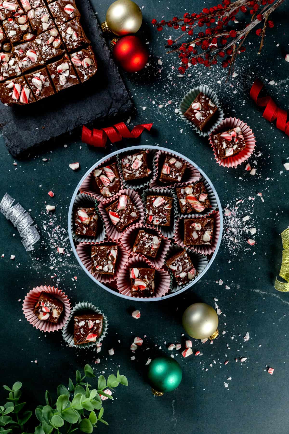 Bites of peppermint fudge with crushed candy canes on top are in a metal tin with more bites next to the tin and on a black serving tray next to it. Christmas ornaments and ribbons are next to the tin.
