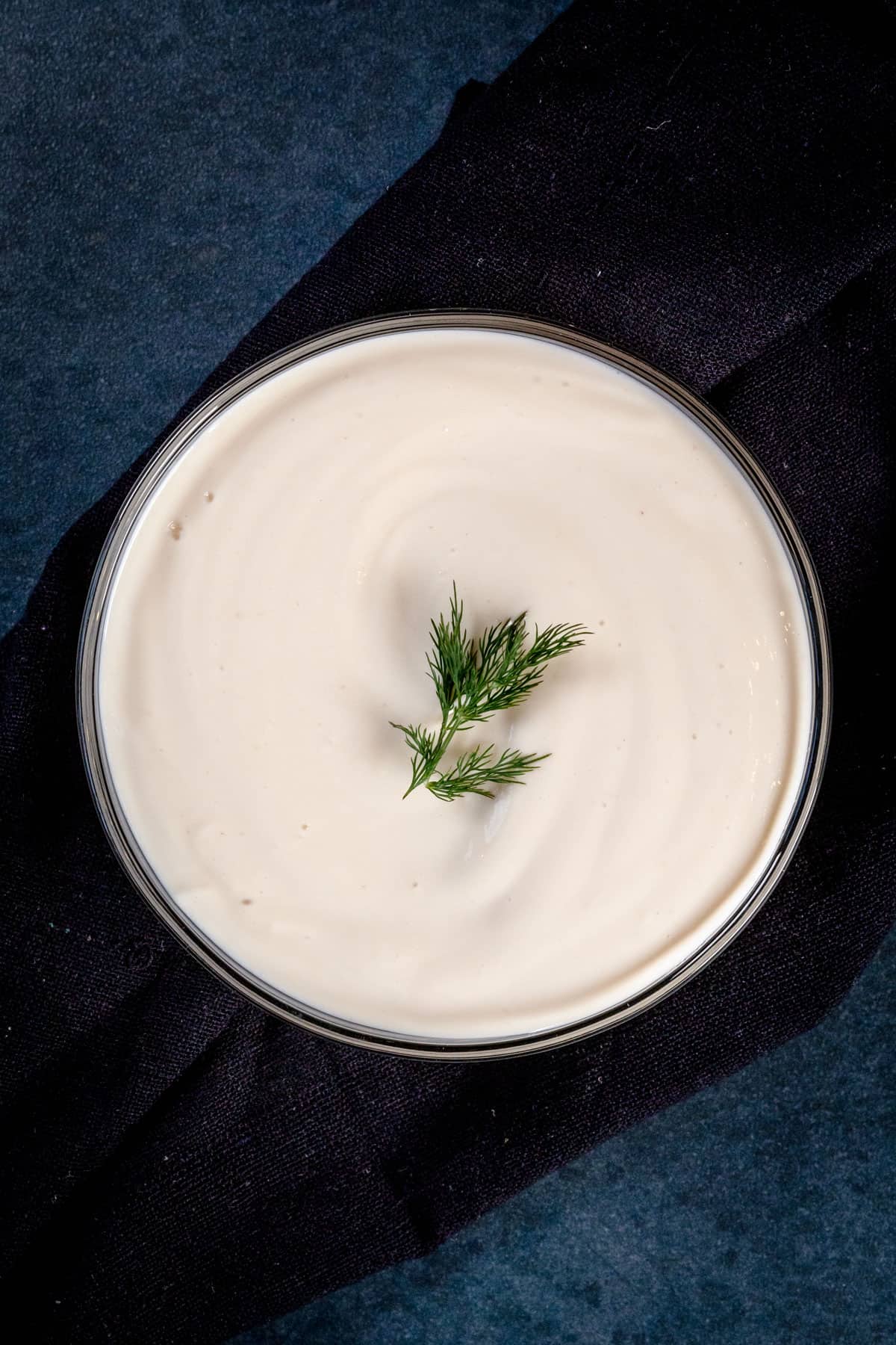 A glass bowl filled with vegan sour cream with a sprig of dill on top of a dark towel.