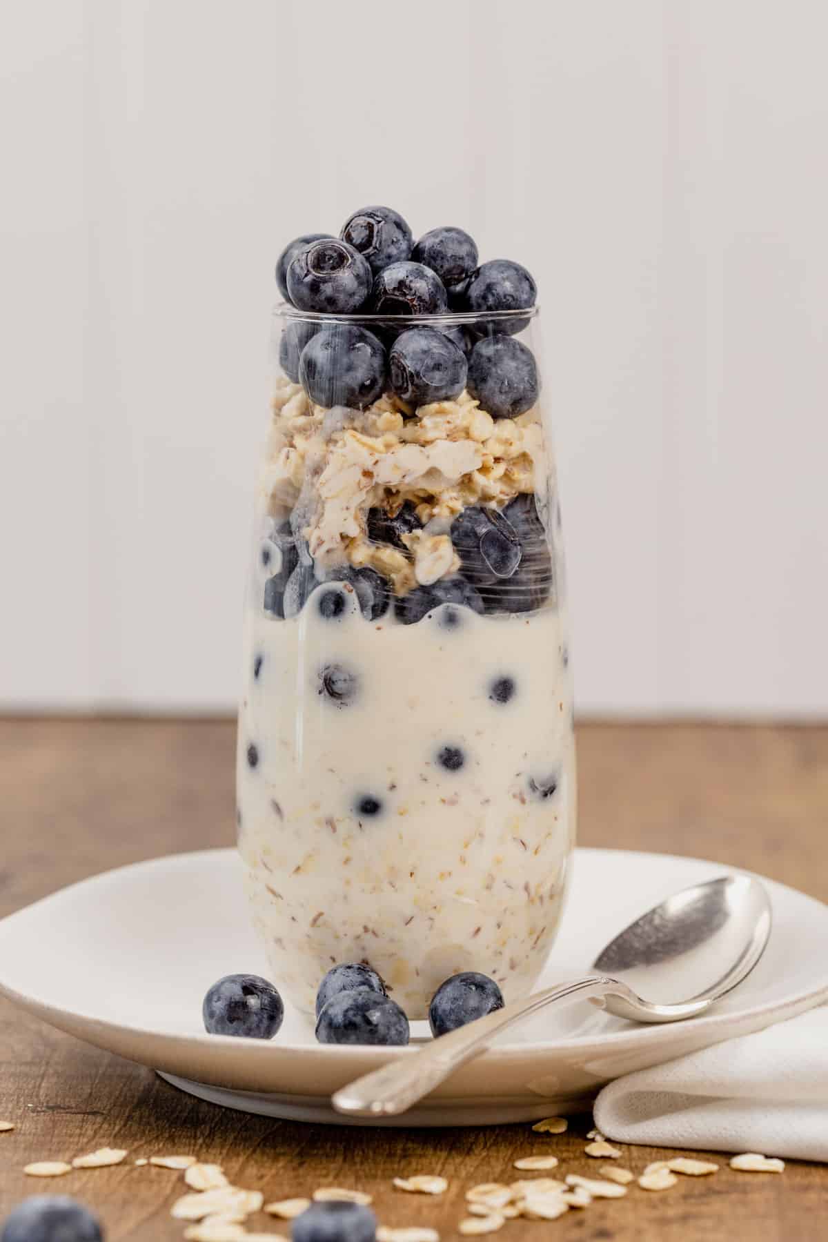A tall glass of vegan blueberry overnight oats on a white plate with a small spoon. Fresh blueberries and oats are surrounding the cup.
