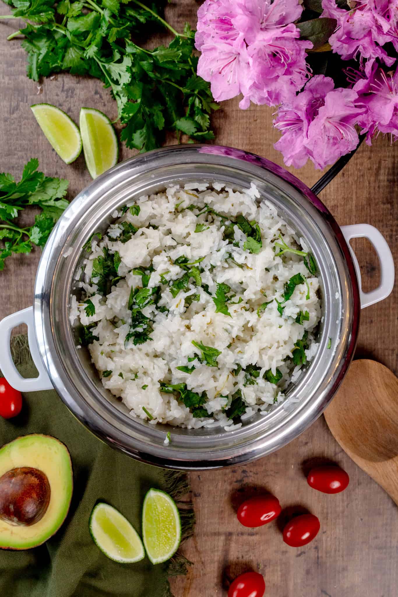 The finished cilantro lime rice in a rice cooker on a wood table surrounded by fresh avocados, limes, tomatoes, cilantro, and flowers.