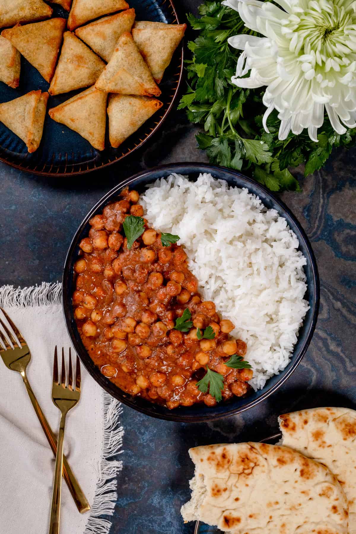 A big blue bowl filled with half white rice and half vegan chickpea curry is on the table surrounded by samosas and naan bread.