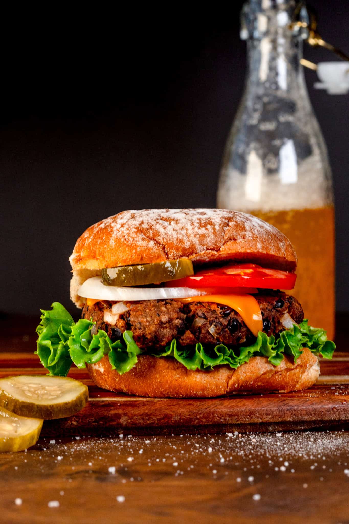 A single black bean burger with lettuce, pickles, tomato, onion, and a cheese slice is on a wood serving tray. Next to the burger are pickles. A glass bottle filled with a fizzy drink is behind the burger.