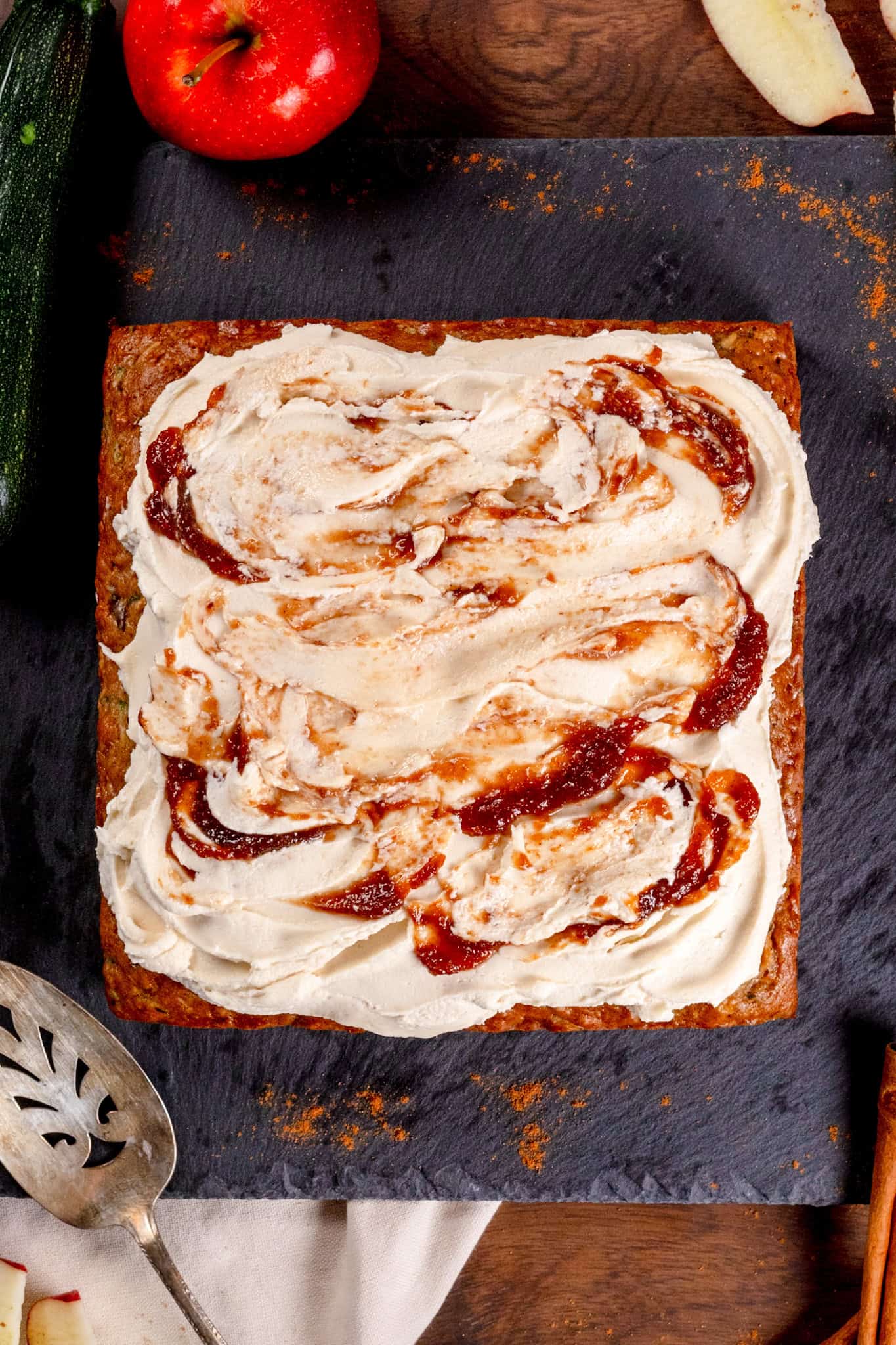 An apple zucchini cake with vanilla and apple butter frosting on a black serving tray with apples and cinnamon sprinkled around.