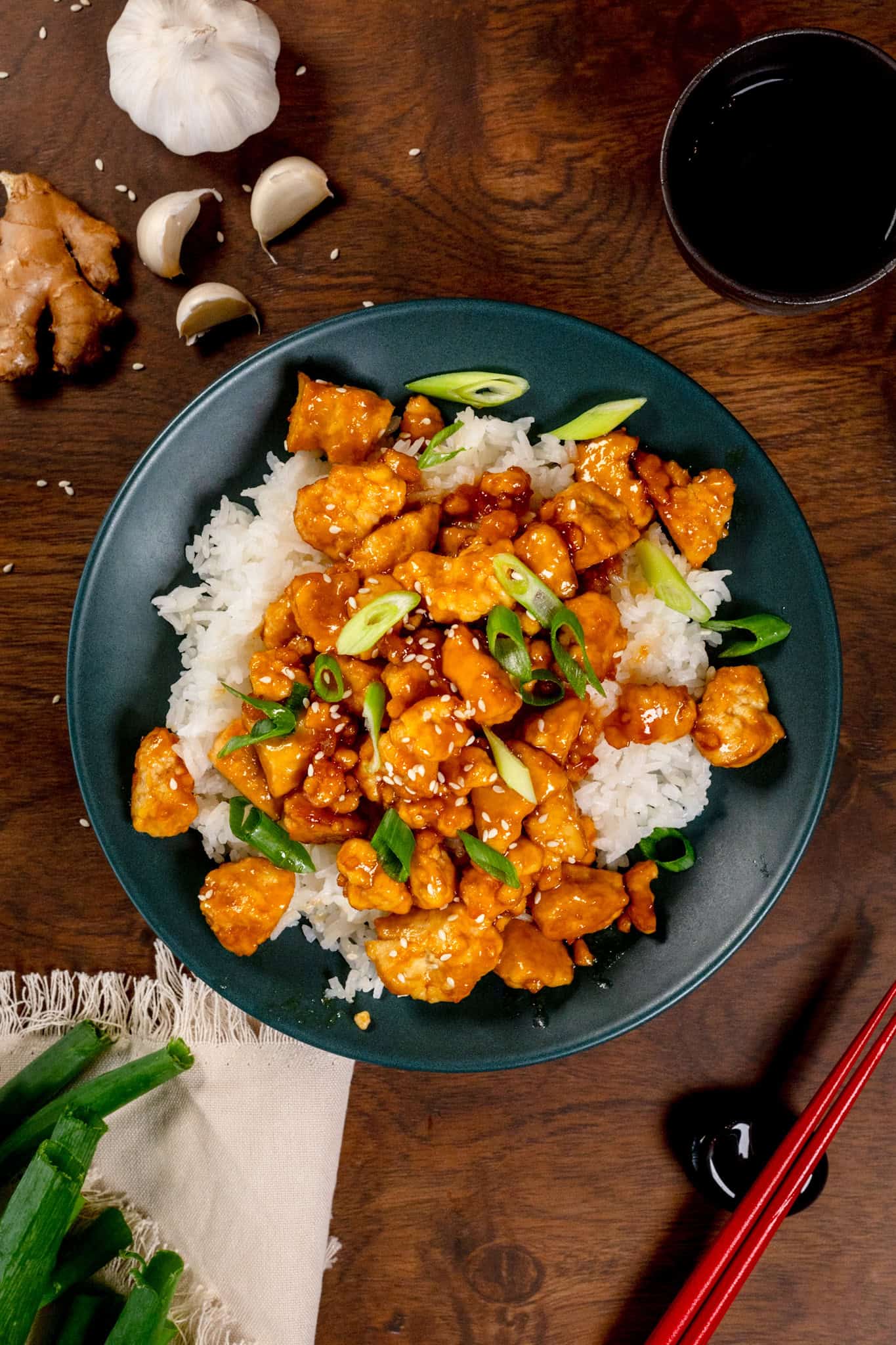 A big plate filled with white rice and topped with pan fried apricot tofu. Fresh green onions are sprinkled on top and ginger and garlic are next to the plate.