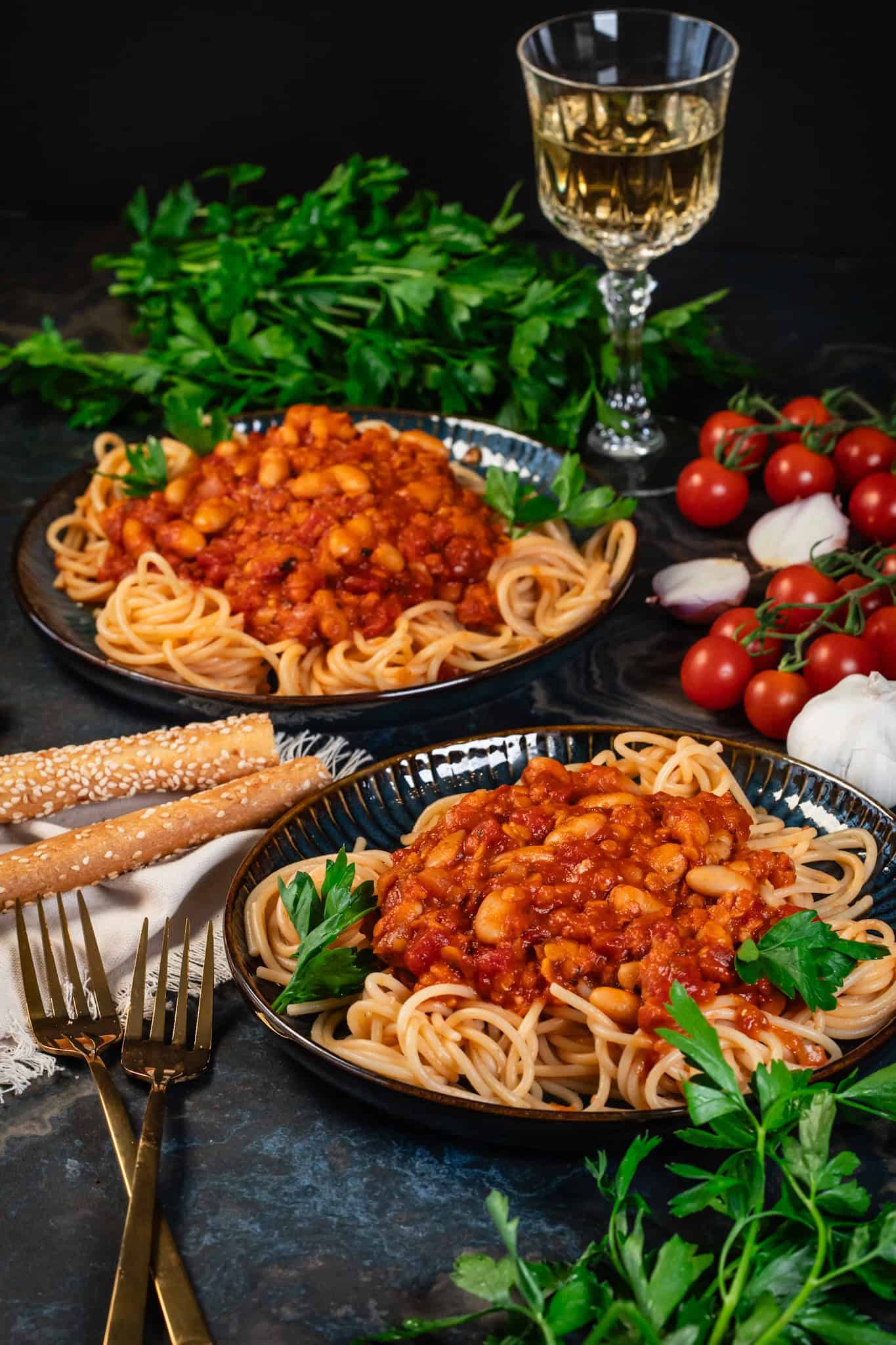 Side view of two dark blue plates filled with spaghetti, protein pasta sauce, fresh tomatoes and parsley, and a few breadsticks.