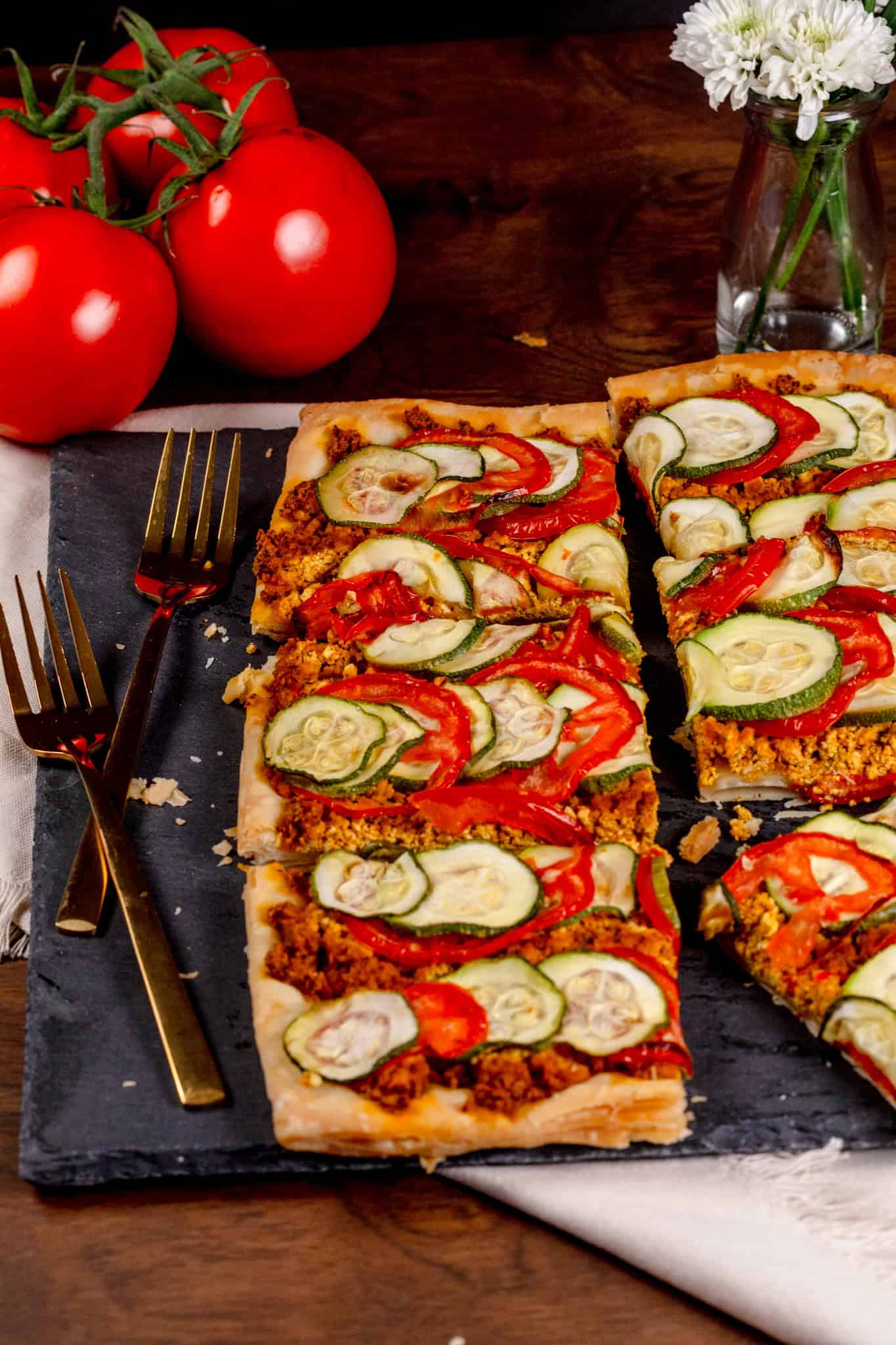The finished tomato and zucchini tart on a black slate serving tray. Gold forks and fresh tomatoes are next to the tart.