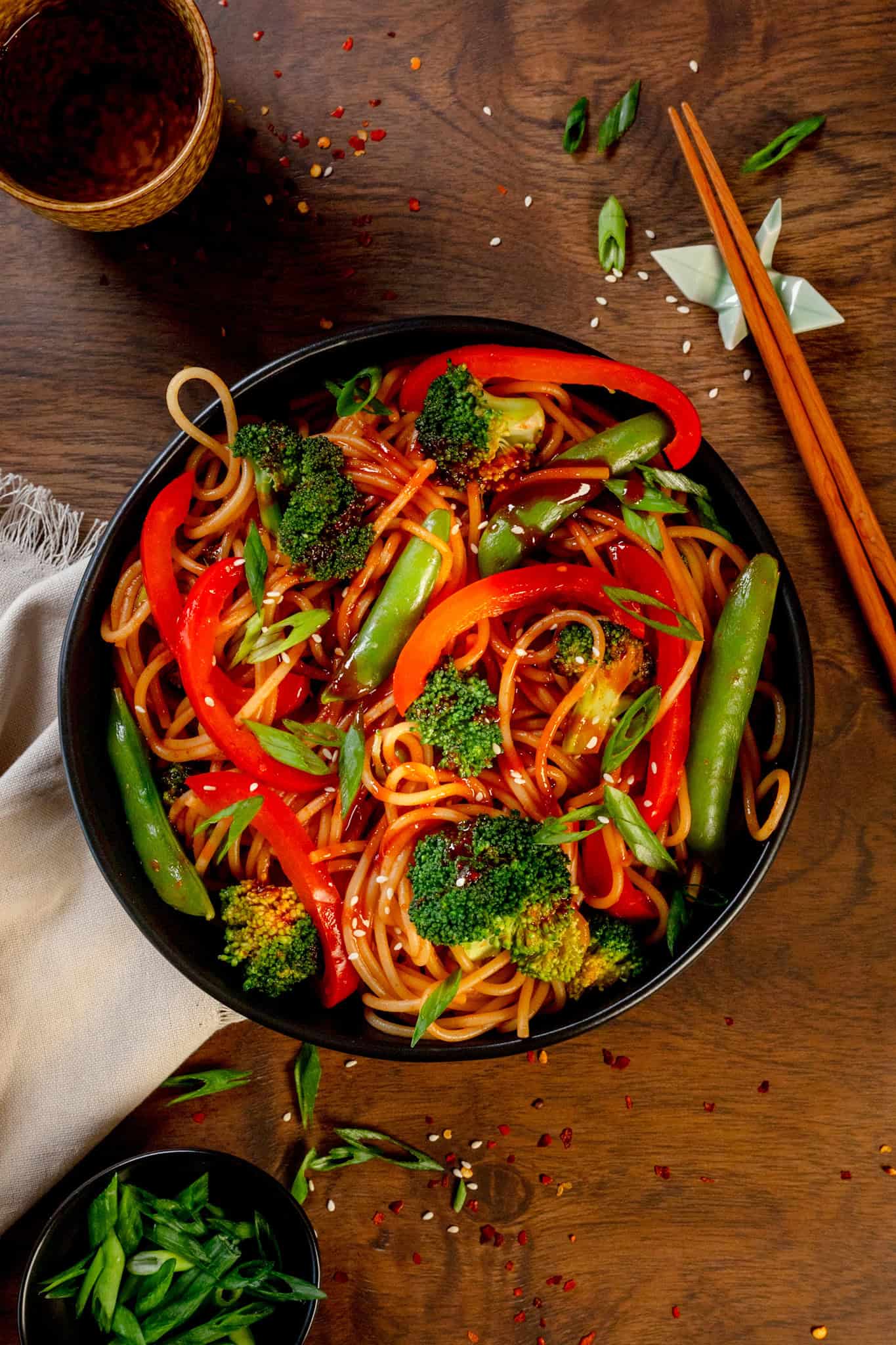 A bowl full of gochujang pasta with lots of stir fry veggies is on a wood table. Chopsticks, extra green onions, sesame seeds, and a glass of water are next to the bowl.
