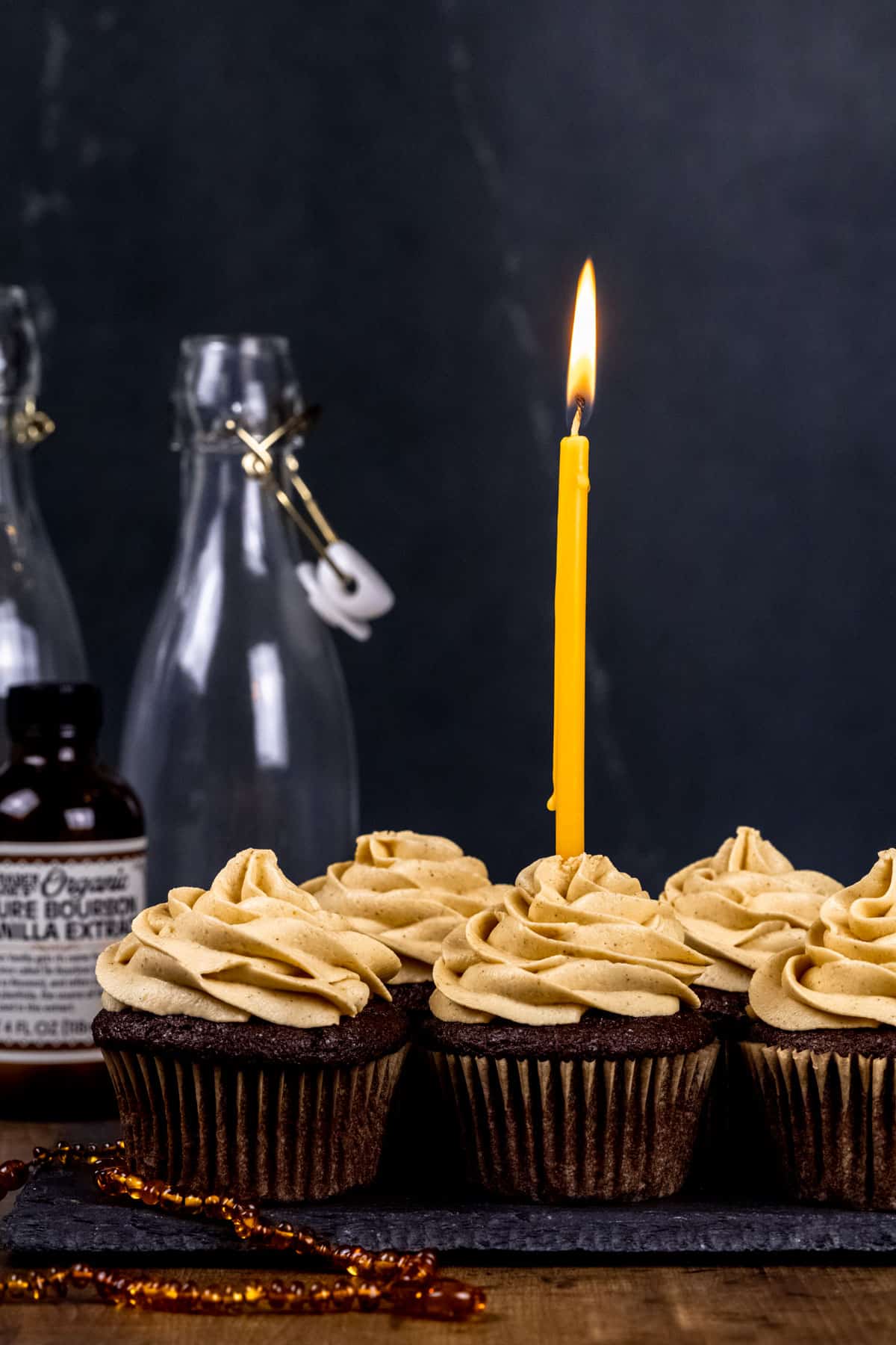 Five chocolate cupcakes with pumpkin buttercream on a table. One cupcake has a long and tall gold candle in the frosting that is lit. Glass bottles are blurred in the background.