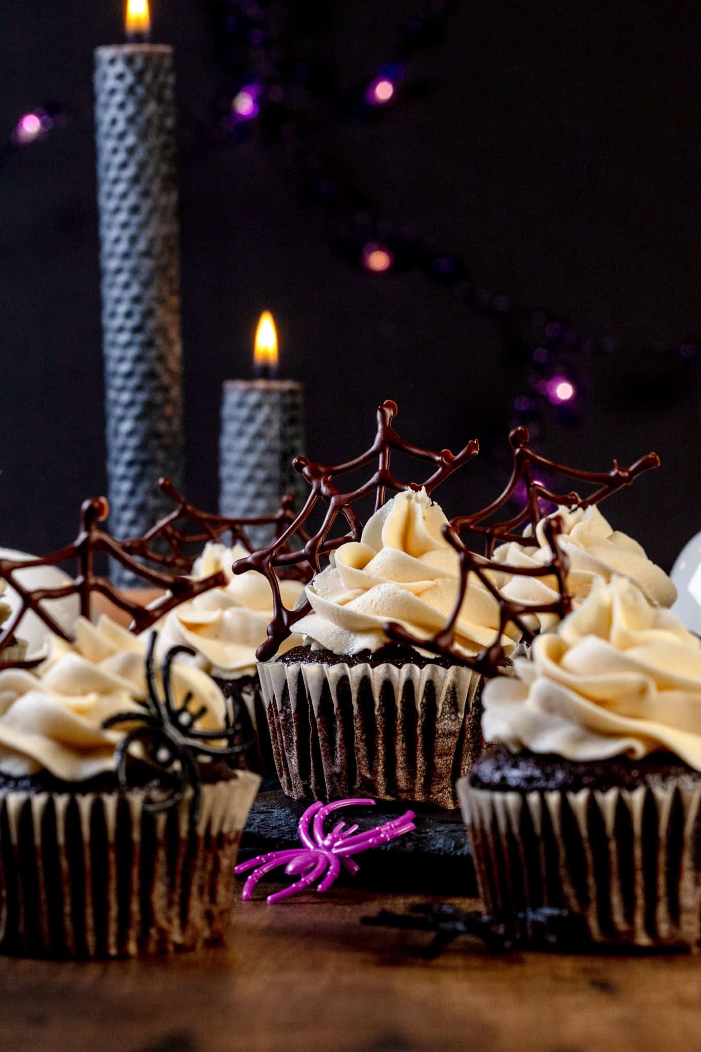Many vegan Halloween spiderweb cupcakes are on a wood table. Spider rings are next to the cupcakes. White frosting is swirled on top and the chocolate spiderwebs are in the frosting. Two black candles are lit in the background. Tiny purple lights are blurred in the background.