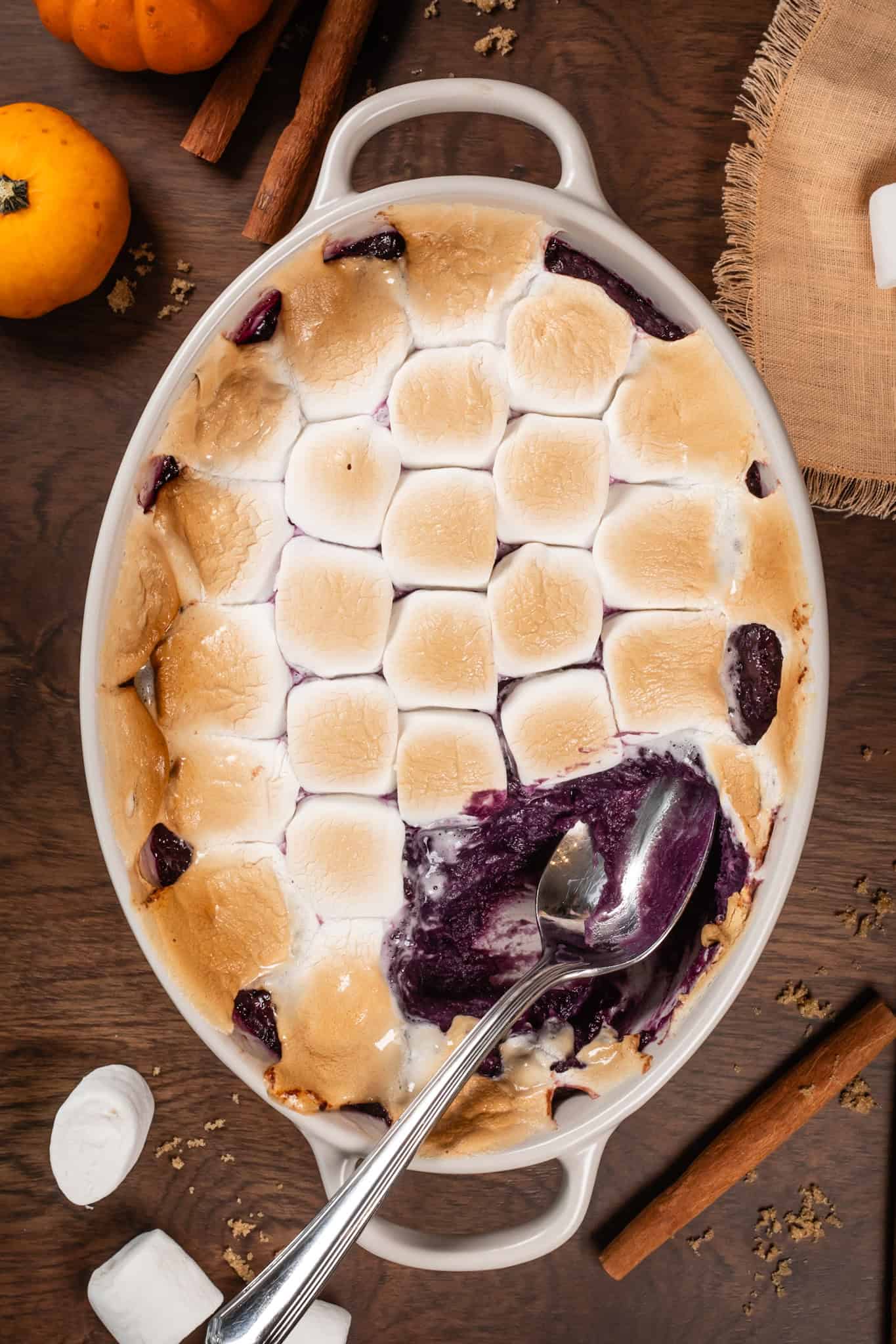 Close up of an oval baking dish filled with vegan purple sweet potato casserole covered in toasted marshmallows. A serving spoon has scooped out a large scoop of the casserole. Mini pumpkins and marshmallows are next to the dish.