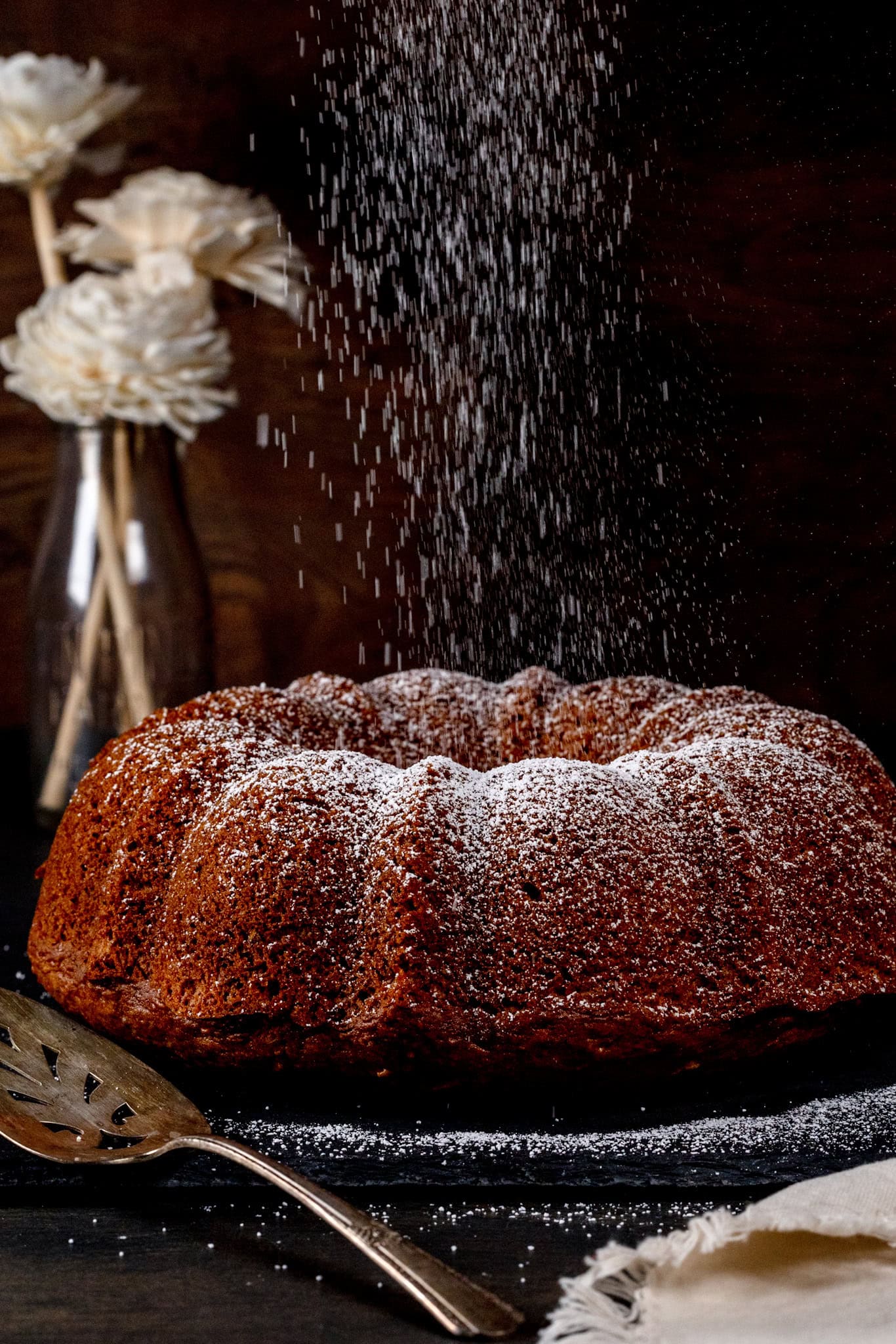 A vegan apple and pear bundt cake is on a dark serving platter next to a serving knife. Powdered sugar is falling like snow on top of the cake. Flowers are blurred in the background.