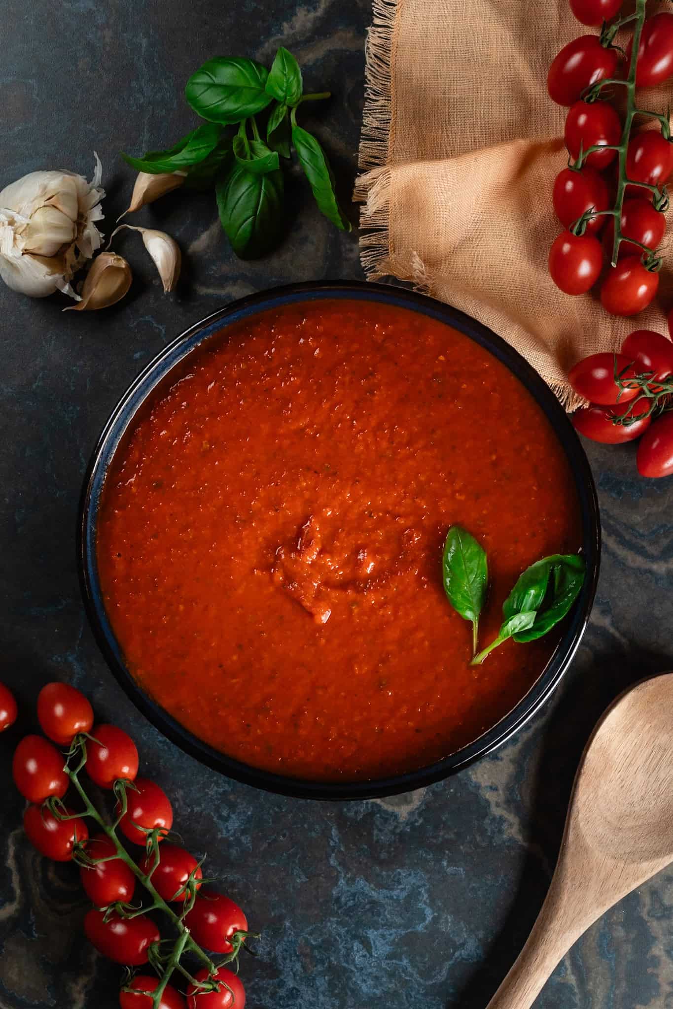 A blue bowl is filled with the finished pizza sauce. Fresh basil and tiny tomatoes are next to the bowl, as well as garlic cloves and a wood spoon. All this sits on a dark blue marble countertop.