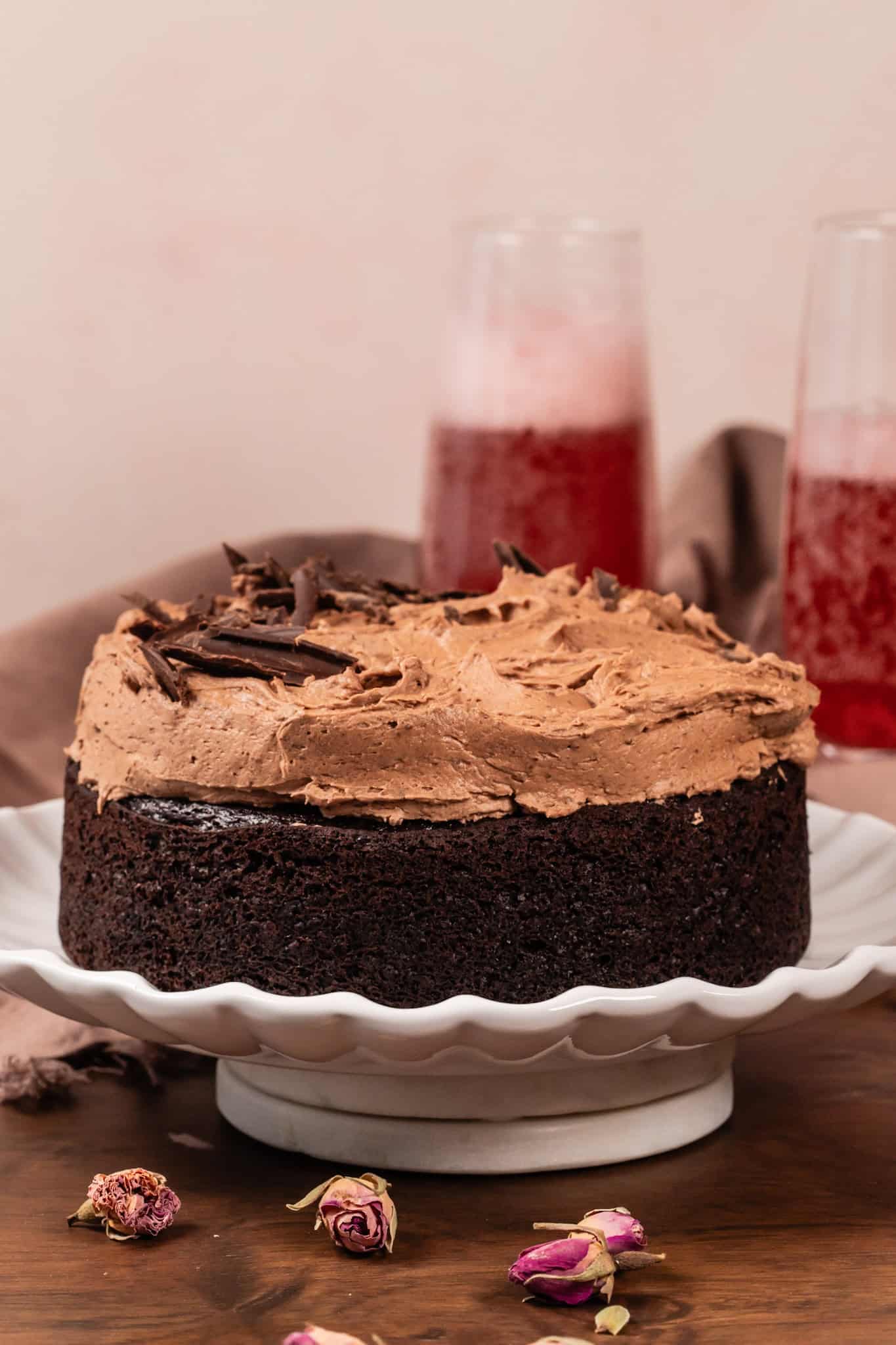 Closeup of a small chocolate cake covered in a thick layer of chocolate buttercream frosting and sprinkled with chocolate shavings. It rests on a white plate on a wood table. Rose buds are next to the cake. Pink fizzy drinks are blurred in the background.