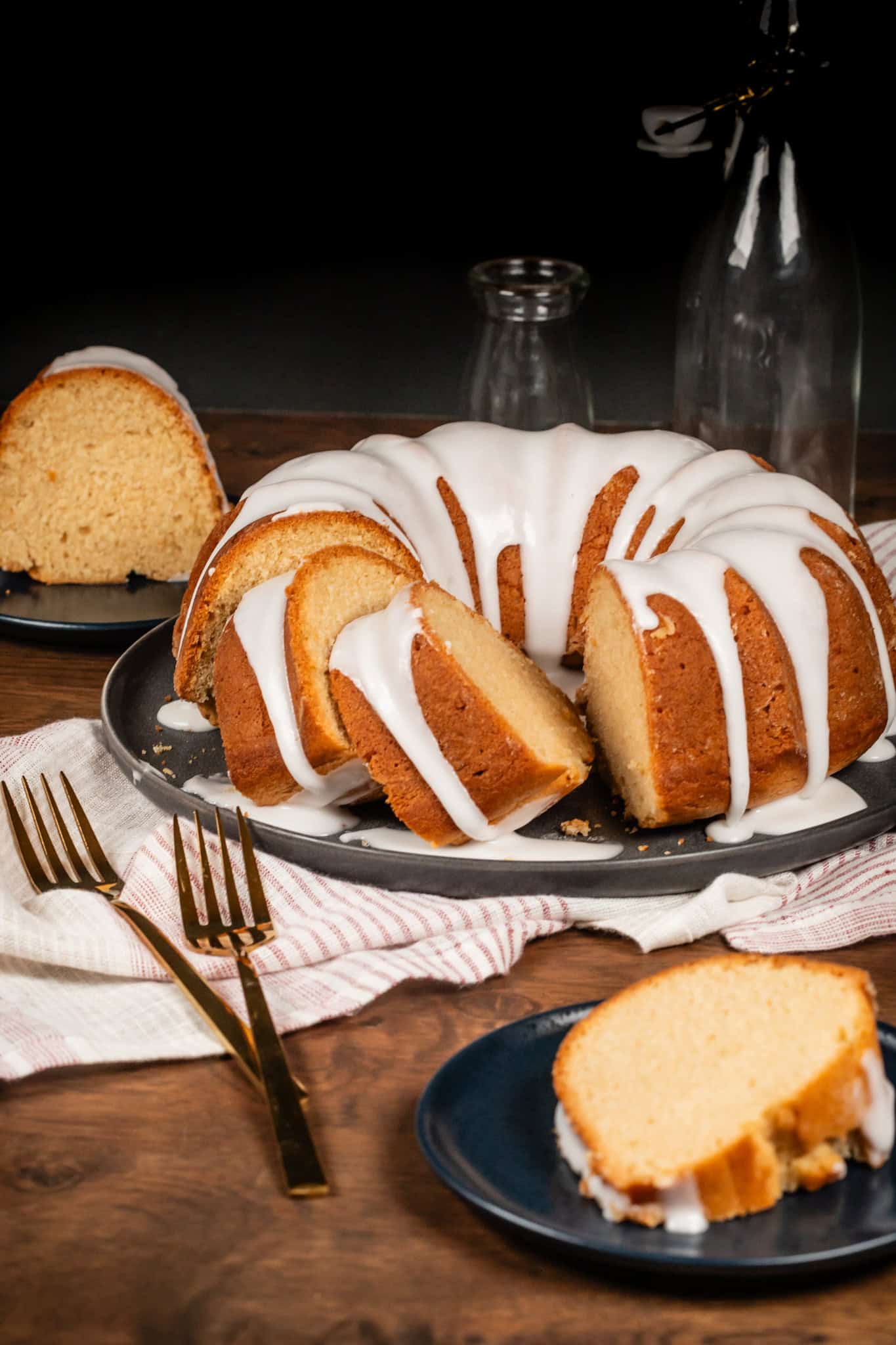 A vegan pound cake in a bundt shape is on a platter. White icing drips down the cake. Slices are cut out and on blue plates in front of and behind the cake. Gold forks are next tot he cake.